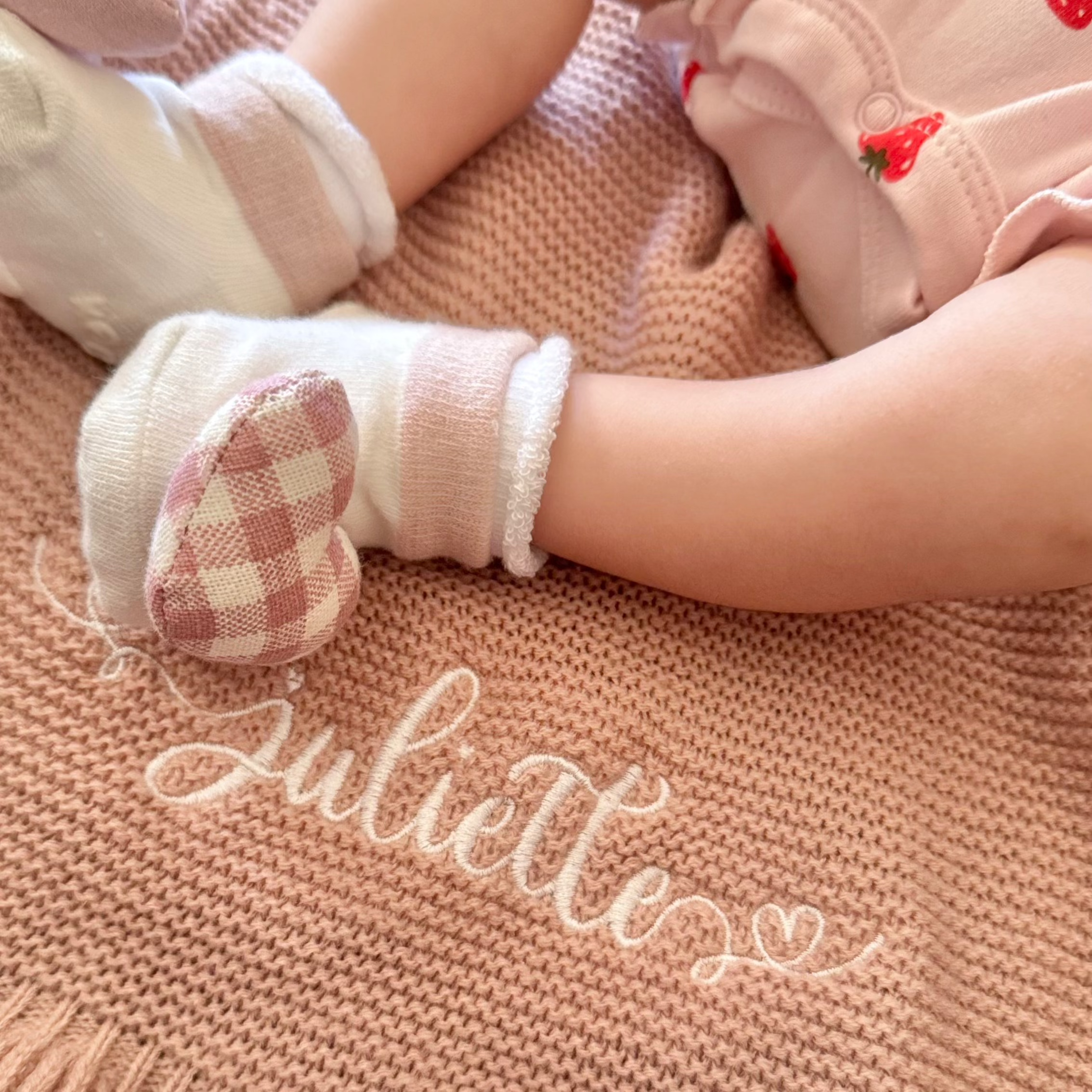 Child's legs wearing white socks with red hearts on a beige blanket with 'Juliette' embroidery.